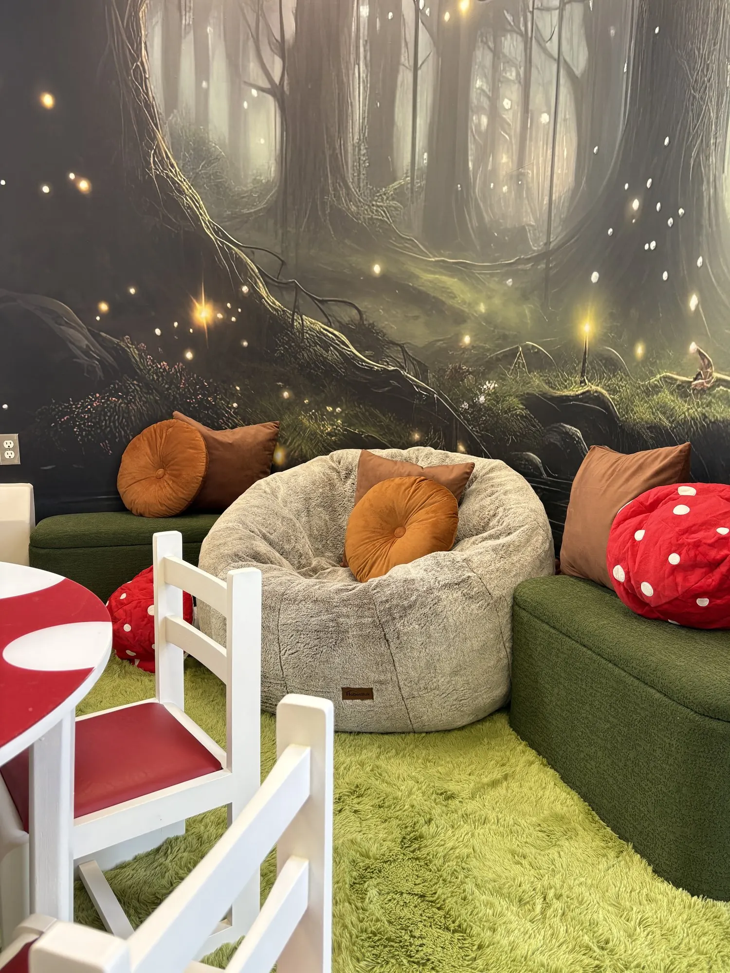 Coin lecture avec pouf, coussins et tapis vert devant une murale de forêt lumineuse