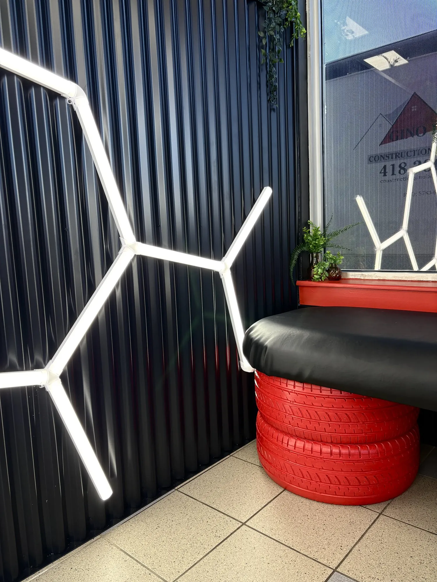 Banquette en pneus rouges et coussin noir au pied du demi-mur en tôle ondulée éclairé au néon